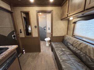 Interior of an Outback LQ horse trailer, featuring a compact living area with a brown leather sofa, wood cabinets, small kitchen counter, and a view into the bathroom with visible toilet. The floor has a stylish wood-patterned finish. -Stillwater Trailer Sales