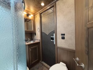 A bathroom inside the Outback LQ Horse Trailer features a glass shower on the left, a sink and mirror in the middle, and a toilet on the right. Wood-toned walls and cabinets complement the frosted glass panel door. -Stillwater Trailer Sales