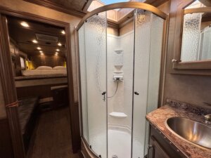 A compact bathroom in an Outback LQ horse trailer features a corner shower with frosted glass doors, a round metal sink on a marble-like countertop, and a view into the Elite 3 Horse bedroom area with bed and seating. -Stillwater Trailer Sales