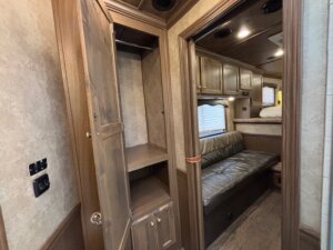 An open wooden closet with shelves is built into the wall of this Outback LQ horse trailer, adjacent to a room featuring a brown leather sofa, overhead cabinets, and a bunk bed in the background. -Stillwater Trailer Sales