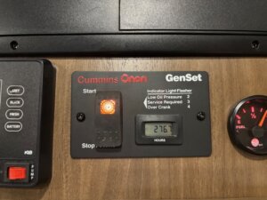 A control panel for a Cummins Onan GenSet inside an Outback LQ Horse Trailer shows an illuminated orange indicator, digital hour meter reading 276.7 hours, and a partial fuel gauge on the right indicating about 1/4 full. -Stillwater Trailer Sales