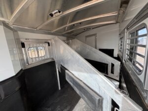 Interior of the Elite 3 Horse Trailer Outback LQ features three angled stalls separated by padded dividers, metal walls, barred windows, and rubber mat flooring, all designed for safely transporting horses. -Stillwater Trailer Sales