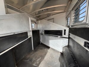Interior view of an empty Elite 3 Horse trailer with metal dividers, black padded walls, and windows with protective bars. The floor appears worn, and sunlight is coming through the windows on the right side. -Stillwater Trailer Sales