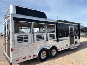 An Elite 3 Horse trailer in black and silver features three side windows, ventilation grills, dual axles, and a rooftop storage area, parked on a gravel lot under a clear sky. -Stillwater Trailer Sales