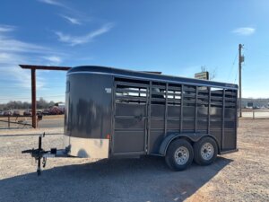 A gray livestock trailer with two axles is parked on gravel under a blue sky. The trailer has a vented upper section and a solid lower section, with a front hitch for towing. -Stillwater Trailer Sales