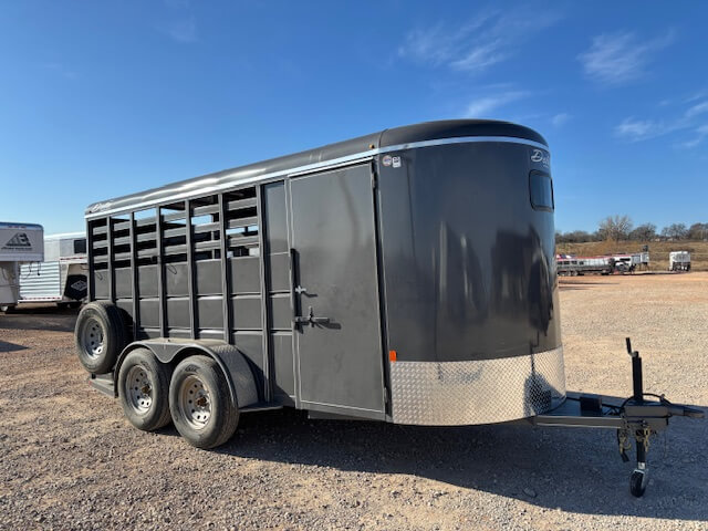 A black, enclosed livestock trailer with ventilation slats, two axles, and a diamond plate front panel is parked on a gravel lot under a clear blue sky. -Stillwater Trailer Sales
