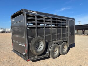 A black Delta livestock trailer with two axles and a spare tire attached to the side, parked on a gravel lot under a clear blue sky. The trailer has open slats for ventilation. -Stillwater Trailer Sales