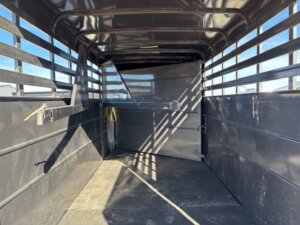 Interior view of an empty black livestock trailer with metal slatted sides, a solid floor, and a closed rear gate. Sunlight is casting shadows inside the trailer. -Stillwater Trailer Sales