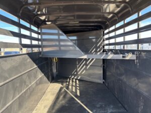 Interior view of an empty livestock trailer with a metal divider and open slatted sides, allowing sunlight and shadows to enter. The floor appears clean and the structure is made of dark metal. -Stillwater Trailer Sales