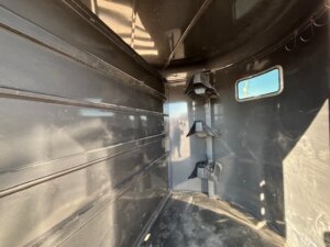 Interior of an empty, black metal trailer with a ribbed wall, small rectangular window on one side, and tiered saddle racks mounted on an upright post. The floor appears dusty and sunlight enters through the window. -Stillwater Trailer Sales