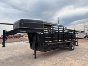 A black Travalong Half Top Stock trailer, 16' long with an open slat design, sits on a gravel lot beneath a cloudy sky. It features one visible axle and a hitch ready for connecting to your truck. -Stillwater Trailer Sales