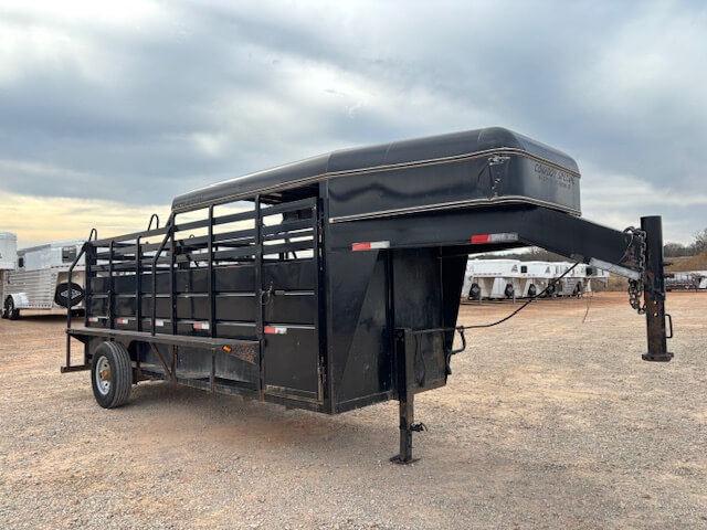 A black Travalong Half Top Stock Trailer with a single axle is parked on a gravel lot under a cloudy sky. Other trailers, including 16' stock trailers, are visible in the background. -Stillwater Trailer Sales