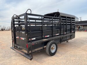 A black Travalong Half Top Stock 16' livestock trailer with an open slatted design is parked on gravel. The trailer has one visible tire, side rails, and a rear gate. Buildings and other trailers are in the background under a cloudy sky. -Stillwater Trailer Sales