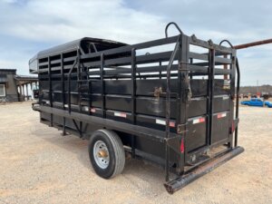 A Travalong 16' Stock Trailer with a half top stock design is parked on a gravel lot. This black, single axle livestock trailer features an open slatted build, rear gate closed, and reflective strips along its sides under a partly cloudy sky. -Stillwater Trailer Sales
