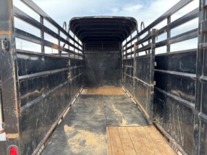 The image shows the inside of an empty black Travalong Half Top Stock trailer with metal bars on the sides, a wooden floor, and an open 16' end facing the viewer. The sky is partly visible through the bars at the far end. -Stillwater Trailer Sales