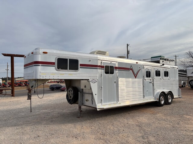 A silver C&C 3 horse trailer with red and black stripes is parked on a gravel lot under a cloudy sky. Featuring living quarters, it has multiple windows, a side door, and two axles with a spare tire mounted in front. -Stillwater Trailer Sales