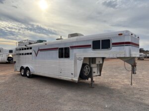 A large white C&C 3 horse trailer with red and gray stripes, featuring living quarters, is parked on a gravel lot under a cloudy sky. The trailer has three axles and a spare tire mounted on the front. -Stillwater Trailer Sales