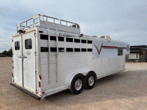 A white C&C gooseneck livestock trailer with ventilation slits, dual axles, a ladder, and rooftop railings is parked on a gravel lot under a cloudy sky. The trailer also features a red stripe and logo detail along its side. -Stillwater Trailer Sales