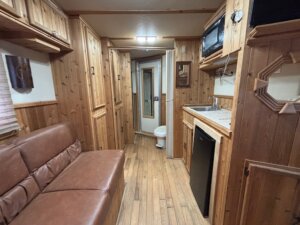 A narrow room with wood paneling serves as the living quarters in this C&C trailer, featuring a brown leather couch, a small kitchenette with sink, fridge, and microwave, plus a door leading to a bathroom with visible toilet. -Stillwater Trailer Sales