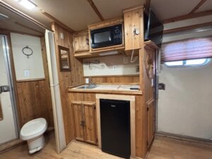 Small Horse LQ kitchenette with wooden cabinets, a sink, black microwave, and mini fridge. A wall-mounted TV is above the counter, and a toilet is visible to the left near a sliding door. A window with a blind is on the right. -Stillwater Trailer Sales