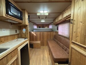 Interior of a wooden-paneled C&C horse trailer with cozy living quarters, featuring a kitchenette on the left, brown leather couch on the right, and a raised sleeping area at the back with microwave, sink, fridge, and windows with blinds. -Stillwater Trailer Sales