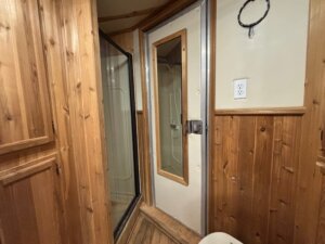A small bathroom in the C&C Horse Trailer features wood-paneled walls, a shower with glass doors, a cream-colored door with a window, an electrical outlet, and a circular wall fixture above the paneling&mdash;perfect for comfortable living quarters. -Stillwater Trailer Sales