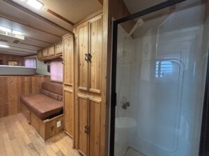 An RV interior in a C&C trailer features wood-paneled walls, cabinets, and floors. A brown cushioned bench sits by a window, with overhead storage above. On the right, the living quarters horse trailer includes a clear glass shower with a white interior. -Stillwater Trailer Sales