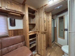 A small C&C 3 horse trailer room with wood-paneled walls and cabinets, open shelving, a brown leather bench, a shower with a glass door, and a toilet. The wood floor and ceiling fixtures add warmth and functionality to this Horse LQ space. -Stillwater Trailer Sales
