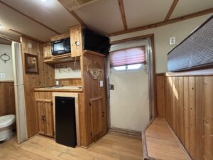 A small kitchenette area in a wood-paneled Horse LQ with a mini fridge, microwave, cabinets, and a door with a window covered by a shade. A bench lines the right wall; a bathroom is partially visible to the left in this C&C 3 horse trailer. -Stillwater Trailer Sales