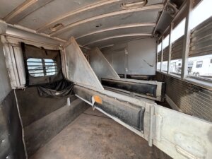 Interior of an empty C&C 3 horse trailer with individual stalls separated by metal dividers, padded sides, and windows on the right letting in light. The floor is bare and slightly dirty&mdash;no living quarters are present. -Stillwater Trailer Sales