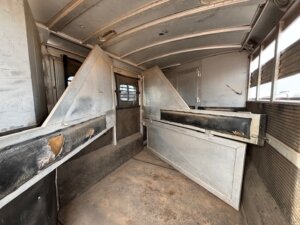 Interior of an empty C&C 3 horse trailer with metal partitions, worn surfaces, and a dirty floor. Sunlight enters through slats on the right side. The dividers and walls show signs of heavy use. -Stillwater Trailer Sales