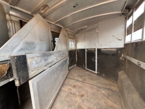 The interior of an empty horse trailer shows metal dividers, a dirty floor, ribbed wall panels, small windows, and a closed door at the far end. Light streams through the windows, illuminating this C&C trailer&rsquo;s spacious layout. -Stillwater Trailer Sales