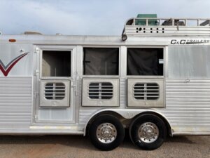 A silver C&C horse trailer with two barred windows and an open door is parked on gravel. The C&C Trailers branding is visible on the right side, set against a partly cloudy sky in the background. -Stillwater Trailer Sales