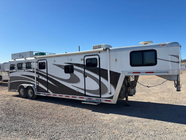 A large white and gray Bloomer 3 Horse gooseneck trailer with living quarters is parked on a gravel lot under a clear blue sky. The 2001 model features multiple windows and decorative swirls on the sides. -Stillwater Trailer Sales