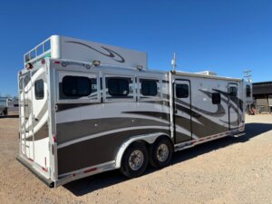 A white and silver 2001 Bloomer gooseneck horse trailer with three windows and two axles is parked on gravel under a clear blue sky. The 3 horse trailer features curved graphic designs and a storage compartment on top. -Stillwater Trailer Sales