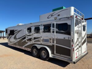 A large, silver and white 2001 Bloomer 3 Horse trailer with multiple windows and two axles is parked on gravel under a clear blue sky. The trailer features decorative graphics and a roof-mounted air conditioning unit. -Stillwater Trailer Sales