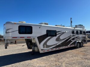 A 2001 Bloomer 3 Horse gooseneck horse trailer in white and gray, featuring four windows, decorative graphics, multiple vents, and a rooftop air conditioner, parked on a gravel lot under a clear blue sky. -Stillwater Trailer Sales