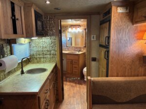 Interior of a 2001 Bloomer 3 Horse RV featuring a small kitchen with sink, wood cabinets, paper towel holder, and a view into the bathroom with vanity, mirror, lights, and toilet. The floor has a stylish wood pattern. -Stillwater Trailer Sales