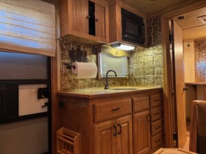 A compact RV kitchen in a 2001 Bloomer 3 Horse trailer features wooden cabinets, countertop, sink with black faucet, mounted paper towel holder, microwave, and a small window with shade. The backsplash displays a decorative metal pattern. -Stillwater Trailer Sales