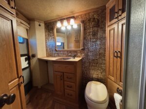 A compact bathroom in a 2001 3 Horse Bloomer features wood cabinets, a sink with storage, a mirror with three lights above, a white toilet, and decorative brown wall tiles. A window and door are visible on the left. -Stillwater Trailer Sales