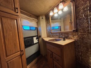 A small bathroom with wooden cabinetry, a single sink vanity, a large mirror with three lights above it, and a closed door with a window covered by a white shade sits inside this 2001 Bloomer 3 Horse trailer. The walls have a metallic embossed pattern. -Stillwater Trailer Sales
