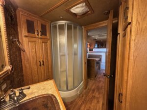 The image shows the interior of a 2001 Bloomer RV bathroom with a curved corner shower, wood cabinets, a sink with a dark faucet, and a view into the living and sleeping area through an open door of this 3 Horse trailer. -Stillwater Trailer Sales