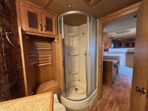 A compact bathroom with a round glass shower enclosure, an open wooden cabinet with shelves and racks, wooden cabinetry, and a view into a living area with wood flooring inside this 2001 Bloomer 3 Horse trailer. -Stillwater Trailer Sales