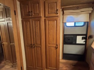 Two tall wooden cabinets stand beside a door with a window and closed shade, echoing the warm tones of the light brown wood floor. A small portion of the countertop is visible&mdash;classic Bloomer 2001 style, reminiscent of a sturdy 3 Horse trailer interior. -Stillwater Trailer Sales