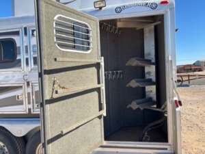 The image shows the open rear tack compartment of a 2001 Bloomer 3 Horse trailer. Inside, there are multiple hooks on the wall and three saddle racks. The trailer is parked on gravel with buildings visible in the background. -Stillwater Trailer Sales