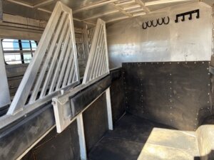 Interior of a 2001 Bloomer 3 Horse livestock trailer with dividers, rubber-lined walls, wall hooks, and sunlight casting shadows on the clean, empty floor. -Stillwater Trailer Sales