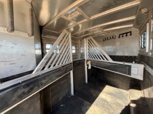 Interior of this 2001 Bloomer 3 horse trailer features metal dividers, padded walls, rubber flooring, wall hooks, and sunlight pouring through side windows. The space is empty, clean, and well-maintained. -Stillwater Trailer Sales