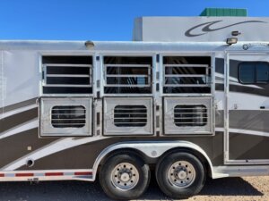 Side view of a 2001 Bloomer 3 Horse trailer with three barred windows, ventilation openings, dual wheels, and a graphic design on the silver and white exterior. The sky is clear and blue. -Stillwater Trailer Sales