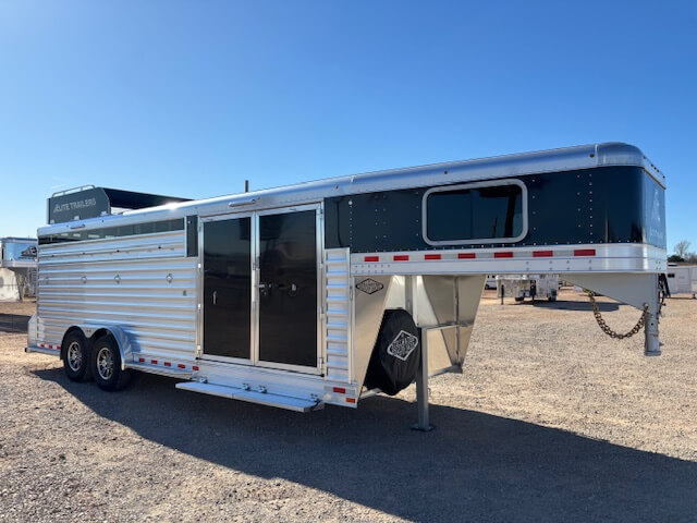 A silver and black Elite 4+1 Combo gooseneck livestock trailer with tandem axles is parked on a gravel lot under a clear blue sky. The trailer features a side door, sliding windows, a Side Tack, and a raised front section for towing. -Stillwater Trailer Sales