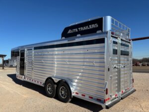 A silver, dual-axle Elite 4+1 Combo livestock trailer with ventilation slats, a rear double-door, and a black sign on top reading ELITE TRAILERS is parked on gravel under a clear blue sky. -Stillwater Trailer Sales